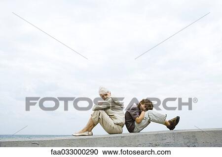 Stock Image - Grandmother and grandson sitting back to back, arms folded, boy looking at camera. Fotosearch