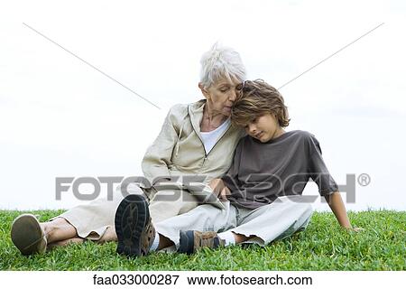 Stock Photo - Grandmother and grandson sitting side by side on the ground, reading book together. Fotosearch