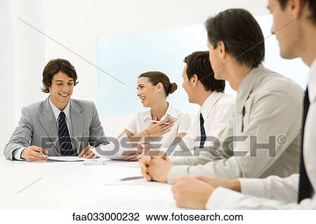 Stock Image - Group of business associates sitting together at conference table, woman smiling at man. Fotosearch