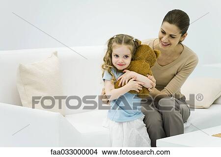 Picture - Little girl holding stuffed bear, woman sitting on couch, gently hugging little girl. Fotosearch