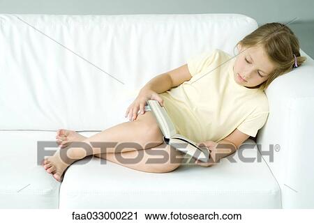 Stock Image - Little girl reclining on sofa, reading book. Fotosearch