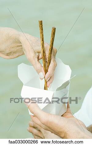 Stock Photograph - Male hand holding container, senior woman's hand reaching in with chopsticks, cropped. Fotosearch