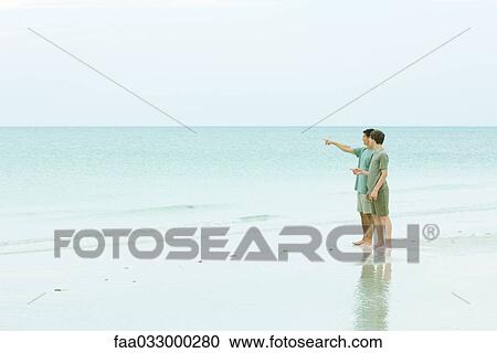 Stock Image - Man and teen boy standing at the beach, looking at view, man pointing. Fotosearch
