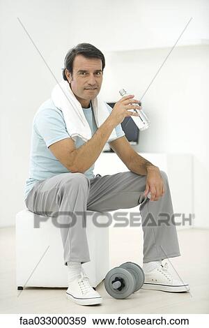 Stock Photo - Man in sports clothing holding water bottle, dumbbell at his feet. Fotosearch
