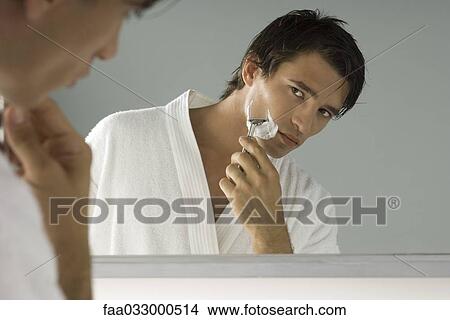 Picture - Man looking at himself in mirror, shaving, wearing bathrobe. Fotosearch