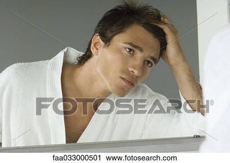 Stock Image - Man looking at himself in mirror, running hand through his hair, wearing bathrobe. Fotosearch