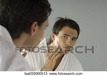 Stock Image - Man looking at himself in mirror, applying shaving cream. Fotosearch
