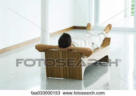 Stock Image - Man lying on bench, hands behind head, rear view. Fotosearch