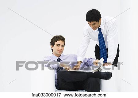 Stock Photo - Man sitting in armchair, newspaper on his lap, smiling at camera, colleague reading over his shoulder. Fotosearch