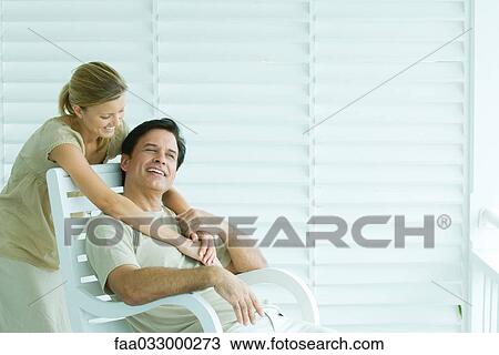 Stock Image - Man sitting in chair, woman standing behind, embracing, smiling. Fotosearch