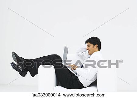 Man slouching in armchair, using laptop, side view View Large Photo Image Picture - Man slouching in armchair, using laptop, side view. Fotosearch