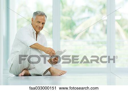 Stock Image - Mature man sitting on the floor, holding book, smiling at camera. Fotosearch
