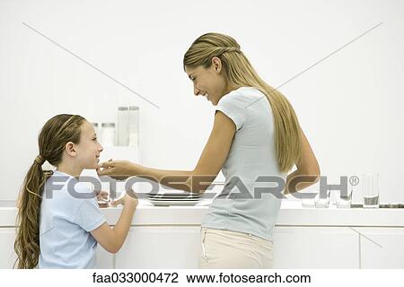 Stock Image - Mother and daughter in kitchen, woman holding girl's chin, both smiling. Fotosearch