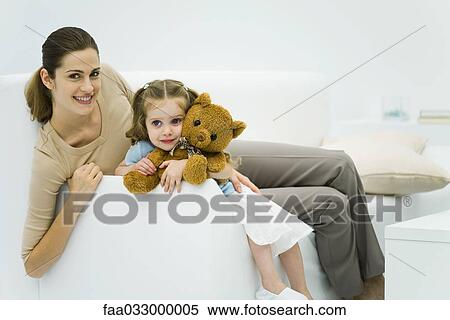 Stock Photography - Mother and daughter sitting on couch, woman smiling at camera, girl holding teddy bear. Fotosearch