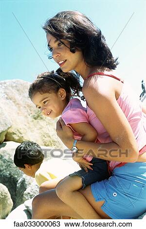 Stock Image - Mother holding daughter on lap outdoors, both smiling, side view. Fotosearch