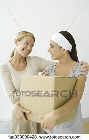 Stock Photo - Mother with arm around daughter's shoulder, daughter holding cardboard box, both smiling. Fotosearch