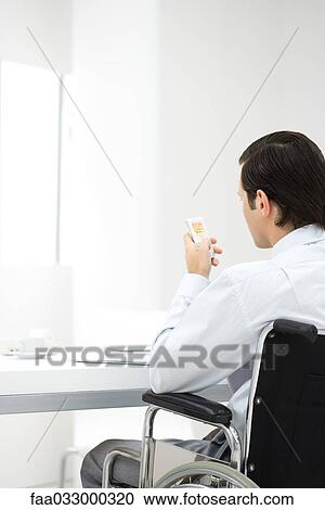 Stock Image - Professional man in wheelchair, looking at cell phone, rear view. Fotosearch