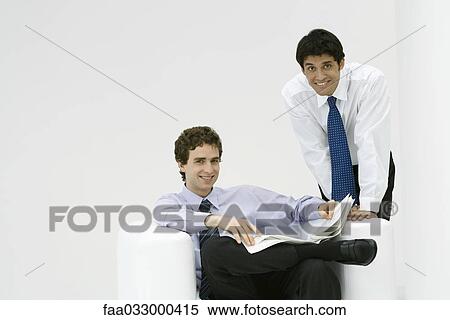Stock Photography - Professional man sitting, holding newspaper, colleague leaning over his shoulder, both smiling at camera. Fotosearch
