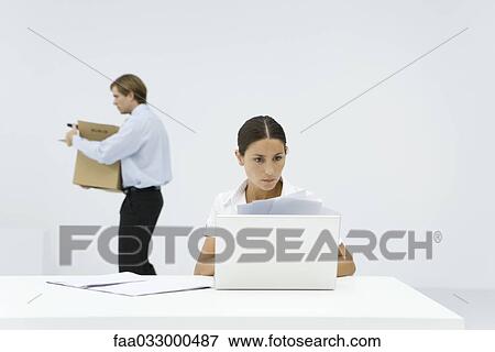 Stock Photo - Professional woman reviewing documents at desk behind laptop, colleague carrying box in background. Fotosearch