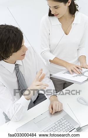 Picture - Professionals discussing graph, female standing and holding clipboard, male sitting and making gesture. Fotosearch