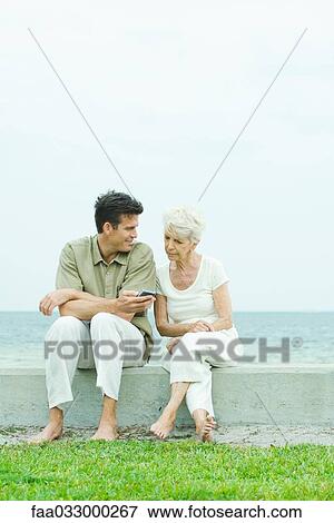 Senior woman and adult son sitting side by side outdoors, man holding cell phone View Large Photo Image Stock Photo - Senior woman and adult son sitting side by side outdoors, man holding cell phone. Fotosearch