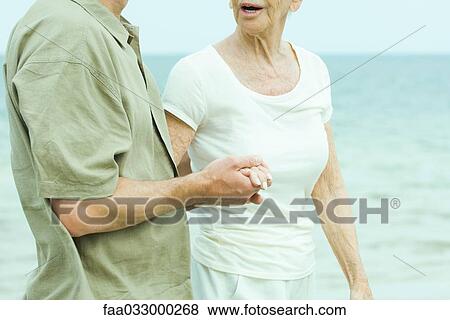 Stock Photo - Senior woman holding adult son's hand, ocean in the background, cropped view. Fotosearch