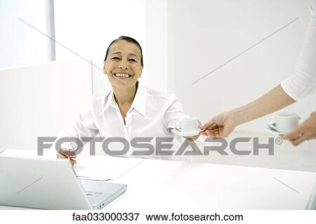 Stock Photo - Senior woman in office, taking coffee from colleague, smiling at camera. Fotosearch