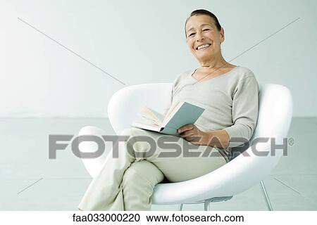 Senior woman sitting in chair, holding book, smiling at camera View Large Photo Image Stock Image - Senior woman sitting in chair, holding book, smiling at camera. Fotosearch