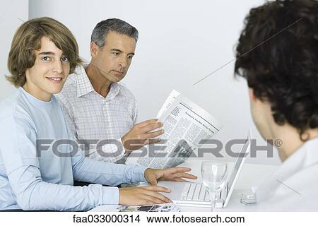 Picture - Teen boy at table with father, using laptop computer, man reading newspaper. Fotosearch