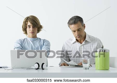 Stock Photography - Teen boy at table with father, using laptop computer, man reading newspaper. Fotosearch