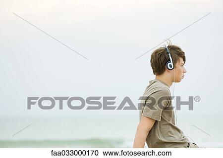 Teen boy sitting on beach listening to headphones, side view View Large Photo Image Stock Image - Teen boy sitting on beach listening to headphones, side view. Fotosearch