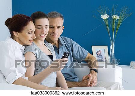 Stock Image - Teen girl on sofa with parents, looking at cell phone together. Fotosearch