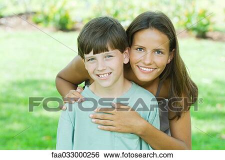Teen girl standing behind younger brother, hands on shoulder and heart, both smiling at camera View Large Photo Image Stock Photograph - Teen girl standing behind younger brother, hands on shoulder and heart, both smiling at camera. Fotosearch