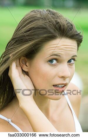 Stock Photograph - Teenage girl holding hand next to ear, looking sideways at camera, close-up. Fotosearch