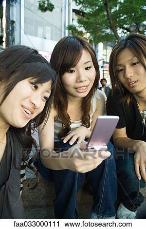 Stock Image - Three young adults looking at cell phone, smiling. Fotosearch