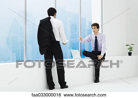 Stock Photo - Two businessmen facing each other, one sitting on ledge, one standing. Fotosearch