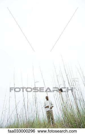 Stock Photograph - Two men standing apart outdoors, low angle view through tall grass. Fotosearch