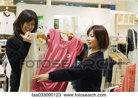 Stock Image - Two young Japanese women in store, holding clothing, one smiling at camera. Fotosearch