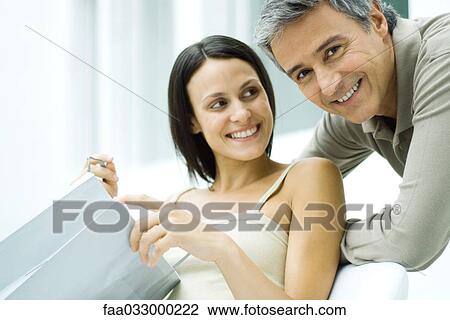 Stock Image - Woman holding gift bag, smiling at man, man smiling at camera. Fotosearch
