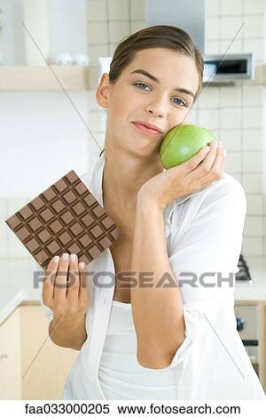 Stock Photography - Woman holding large chocolate bar in one hand, apple in the other, smiling at camera. Fotosearch