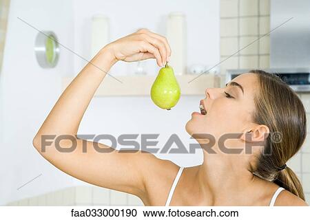 Stock Image - Woman holding up pear, head back, mouth open, side view. Fotosearch