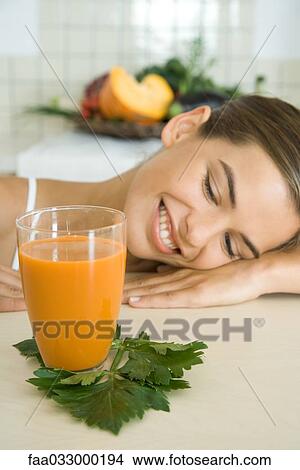 Picture - Woman resting head on counter, looking at glass of vegetable juice, smiling. Fotosearch