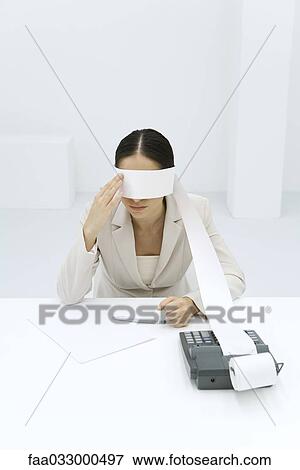 Stock Photo - Woman sitting at desk, tape from an adding machine wrapped around her eyes, touching face. Fotosearch