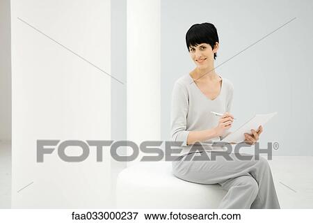 Stock Photo - Woman sitting, holding notepad and pen, smiling at camera. Fotosearch