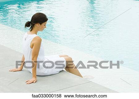 Stock Photo - Woman sitting on edge of pool, dangling feet in water, side view. Fotosearch