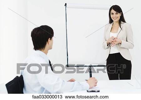 Woman standing in front of presentation board, smiling at camera, male colleague watching View Large Photo Image Stock Image - Woman standing in front of presentation board, smiling at camera, male colleague watching. Fotosearch