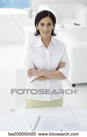 Stock Image - Woman standing with arms folded, smiling at camera, documents on table in foreground. Fotosearch
