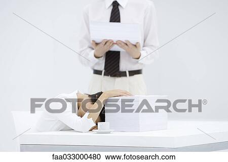 Stock Image - Woman with head down on desk, next to tall stack of papers, man holding another stack of paper. Fotosearch