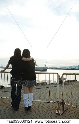 Stock Image - Young couple standing together, looking at waterfront, rear view. Fotosearch