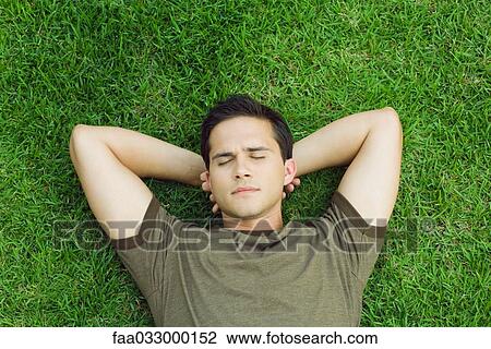 Stock Image - Young man lying on grass with hands behind head, eyes closed, high angle view. Fotosearch
